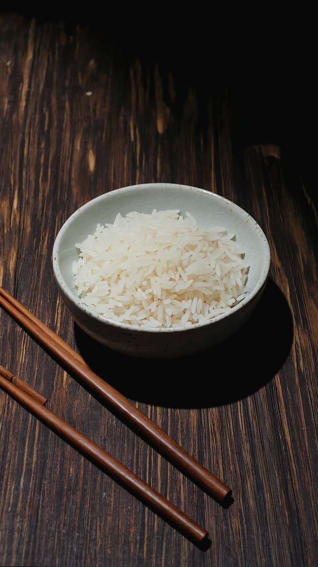 A bowl of white rice with chopsticks laid beside it on dark wood, tense minimal composition, low-key dramatic lighting